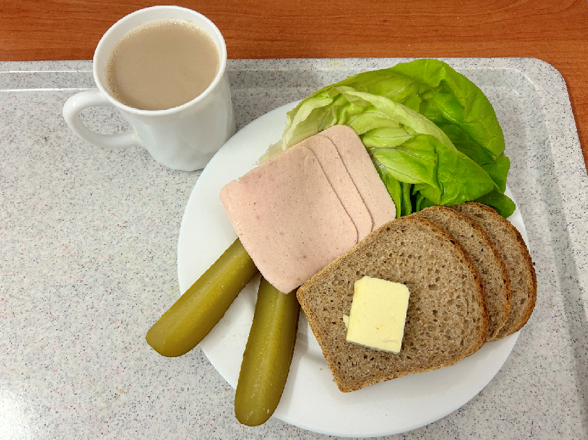 Na zdjęciu znajduje się: Kawa z mlekiem, Chleb Graham, Masło extra 82%, wędlina drobiowa parzona z dodatkiem mięsa wieprzowego, Ogórek kiszony, ​​​​​​​Sałata zielona